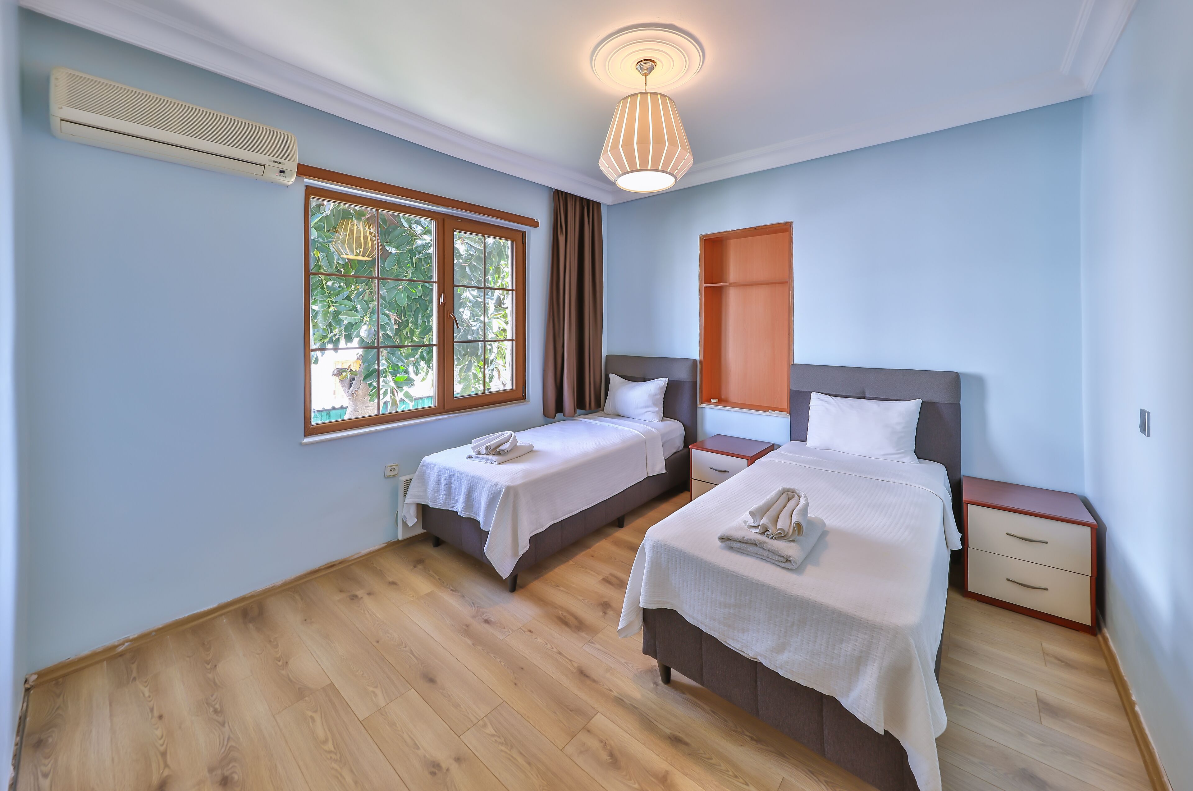 Une chambre d’hôtel avec deux lits, une fenêtre avec des rideaux, un luminaire au plafond et un plancher en bois.