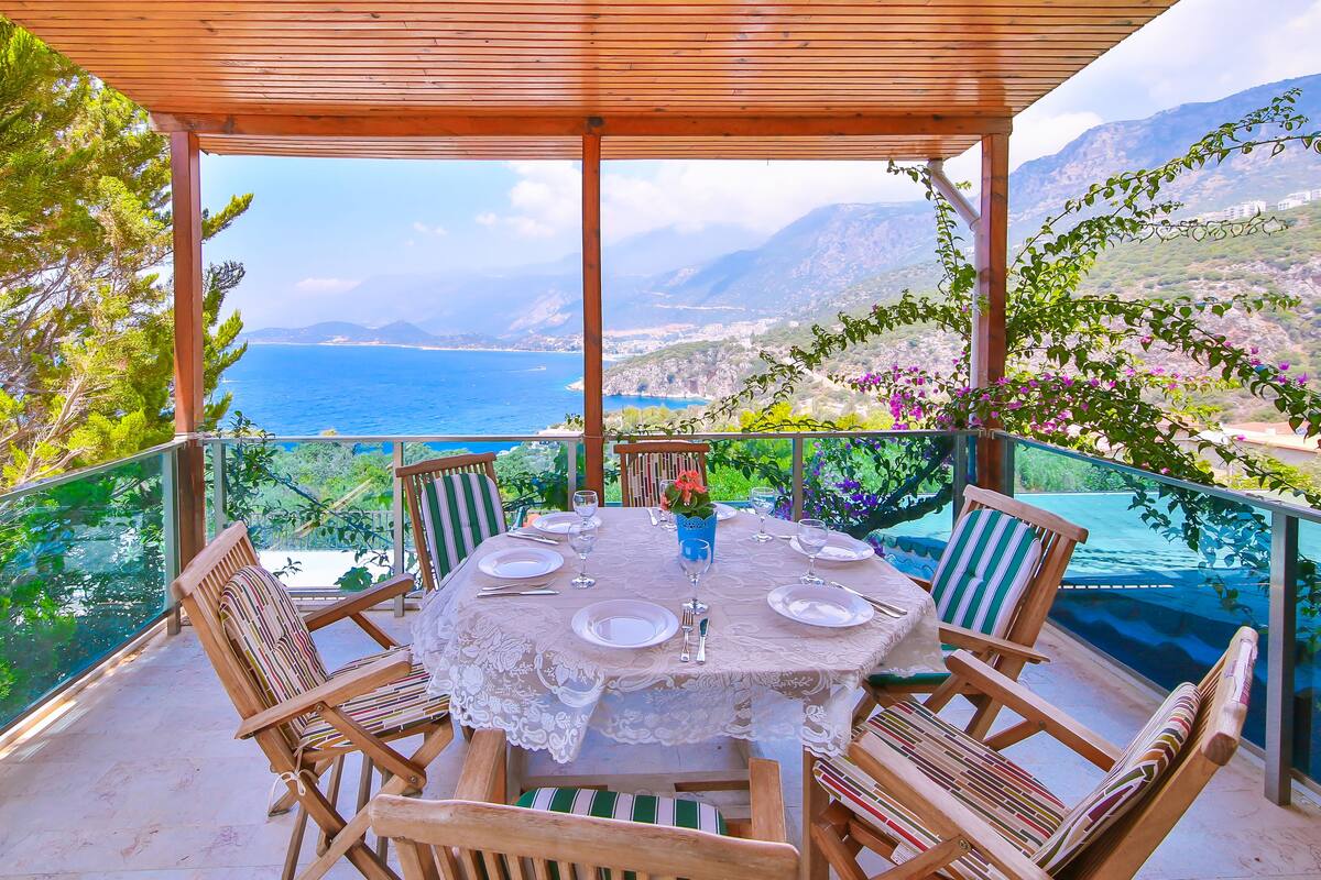 Un área de comedor al aire libre techada con una mesa dispuesta para cuatro personas, con vistas a un paisaje costero y montañas al fondo.