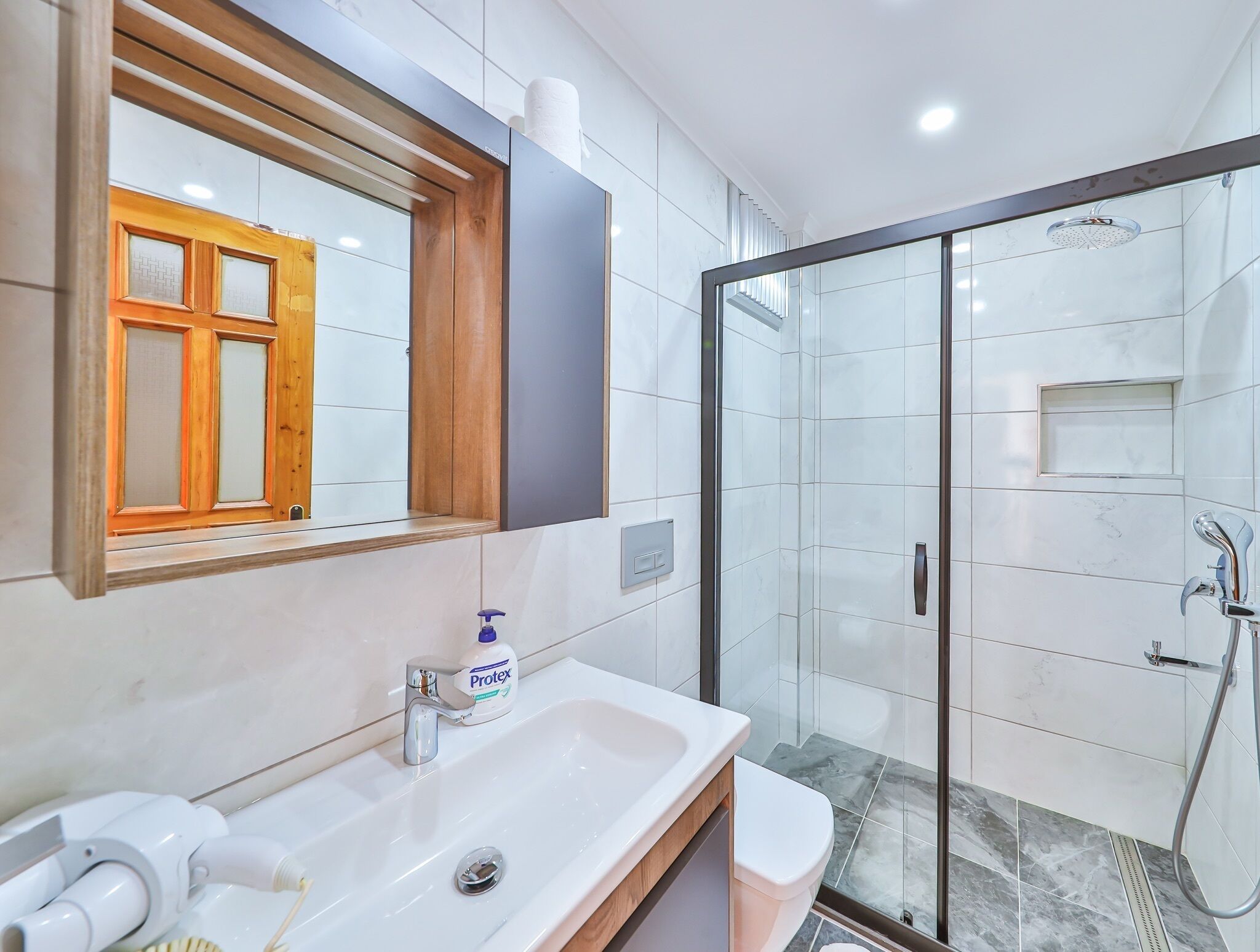 Un baño moderno con lavamanos blanco, tocador de madera, cabina de ducha con mampara de vidrio y espejo con marco de madera.