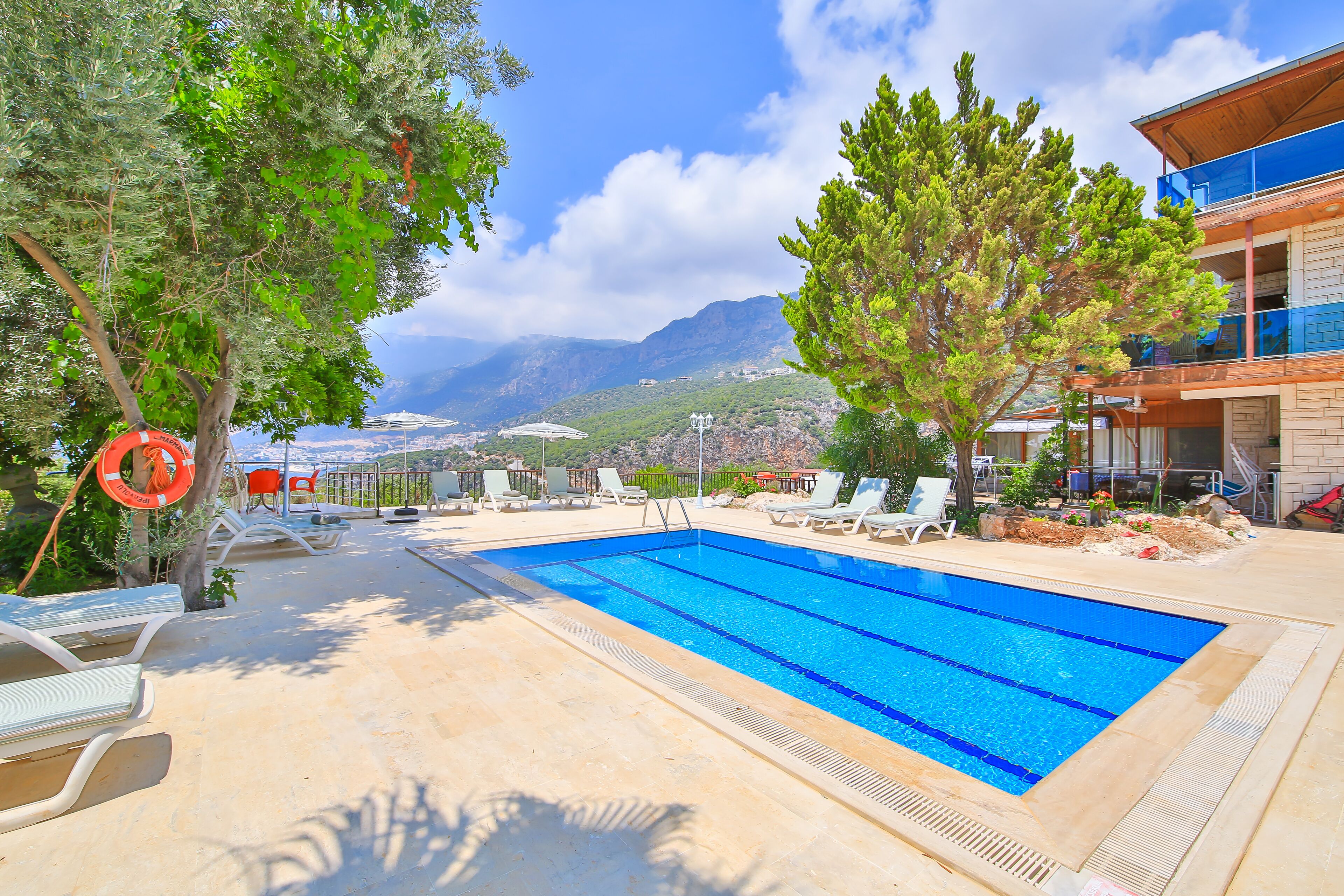 A swimming pool with lounge chairs and a lifebuoy, surrounded by trees and a mountainous backdrop.
