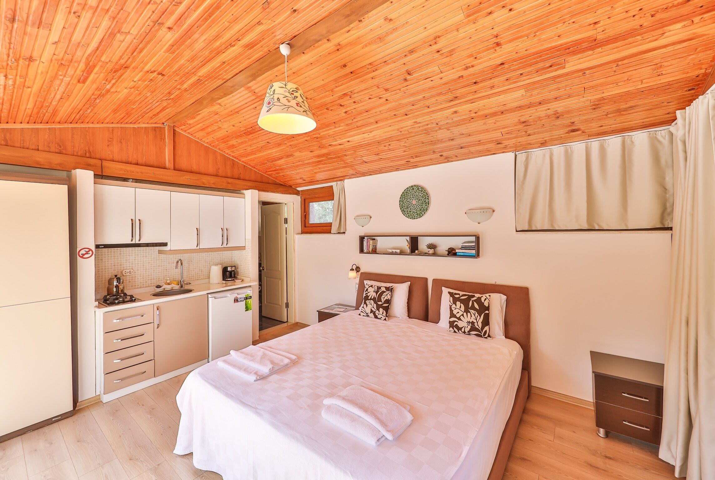 A bedroom with a large bed, a wooden ceiling, a small kitchenette, and a bathroom.
