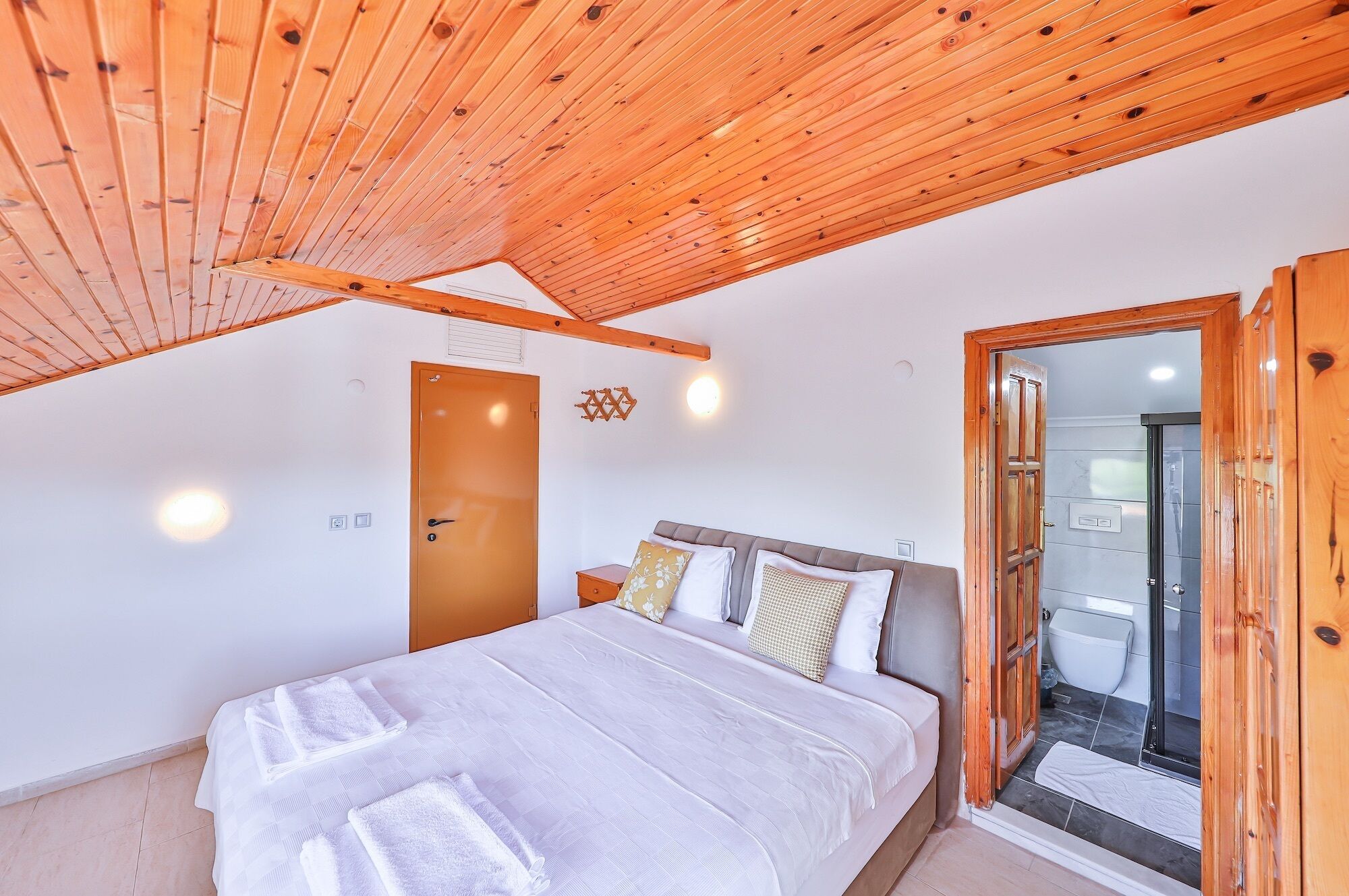 Un dormitorio con techo de madera, una cama con ropa blanca y una puerta, además se ve un baño por una puerta abierta.