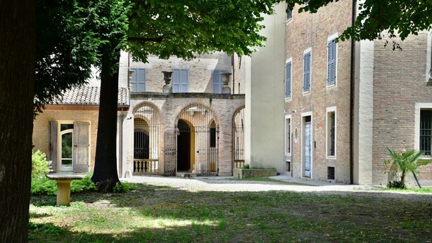 A courtyard with a brick building featuring arched doorways and a large tree providing shade.