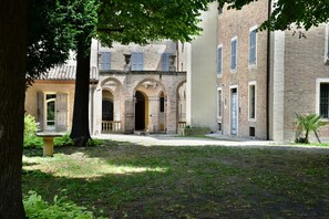 Pátio com um edifício de tijolos, portas em arco e uma grande árvore que oferece sombra.