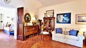 A room with a tiled floor, a white sofa with blue cushions, a wooden cabinet, a chair, a bed with a striped bedspread, and a wall with framed artwork.