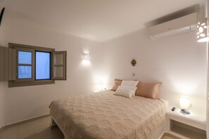A bedroom with a bed, bedside lamps, an air conditioning unit, and a window with shutters.
