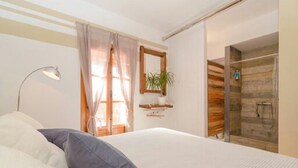Una camera da letto con un letto, una lampada da comodino, una finestra con tende, uno specchio, una pianta e uno spazio doccia.