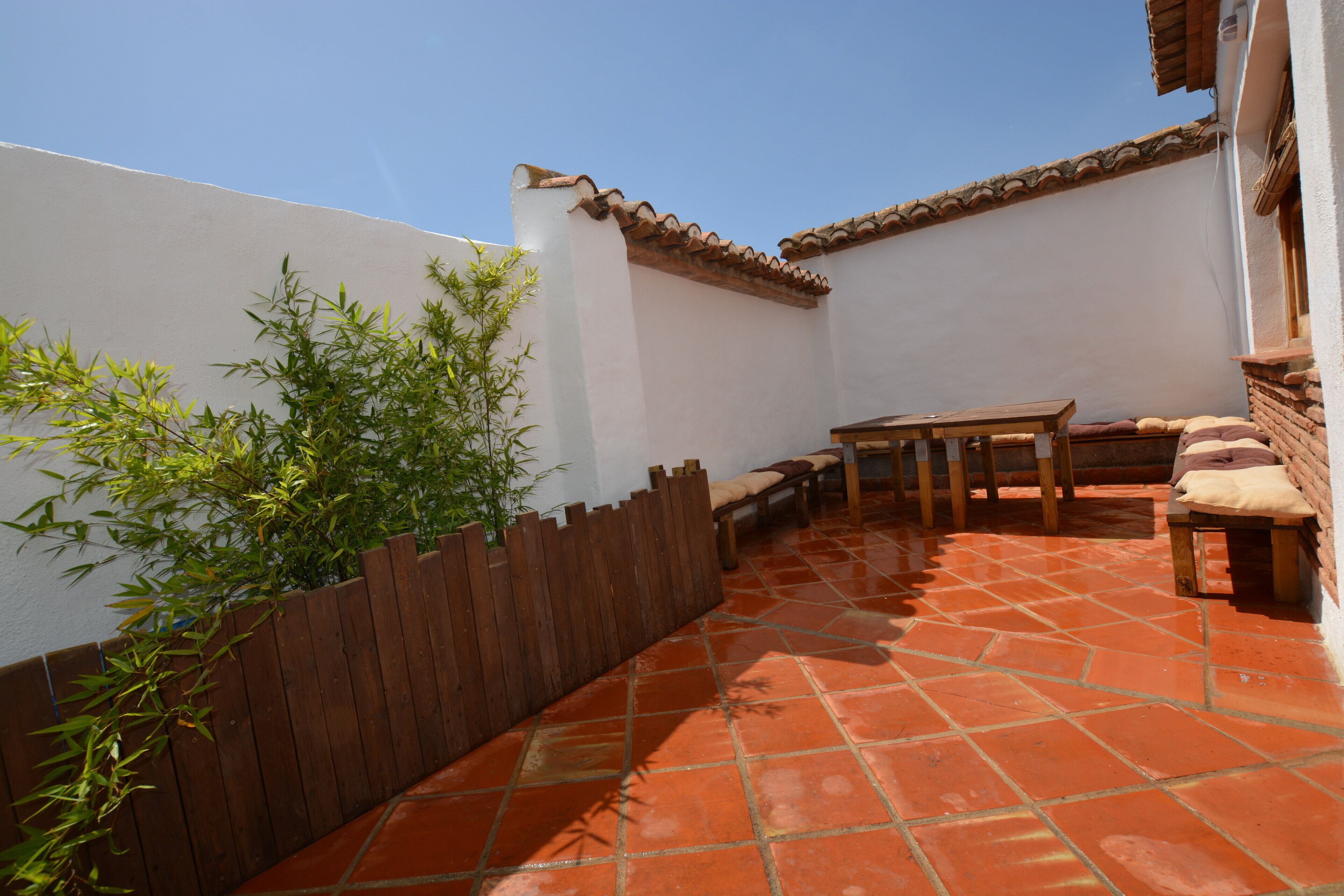 A courtyard with a wooden table and benches, surrounded by white walls and a tiled floor.