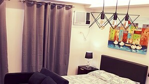 A bedroom with a bed, a nightstand, a lamp, a chair, a table, and a painting on the wall. - BACOLOD CITY CENTER  SLEEK STUDIO DE LUXE (Bacolod)