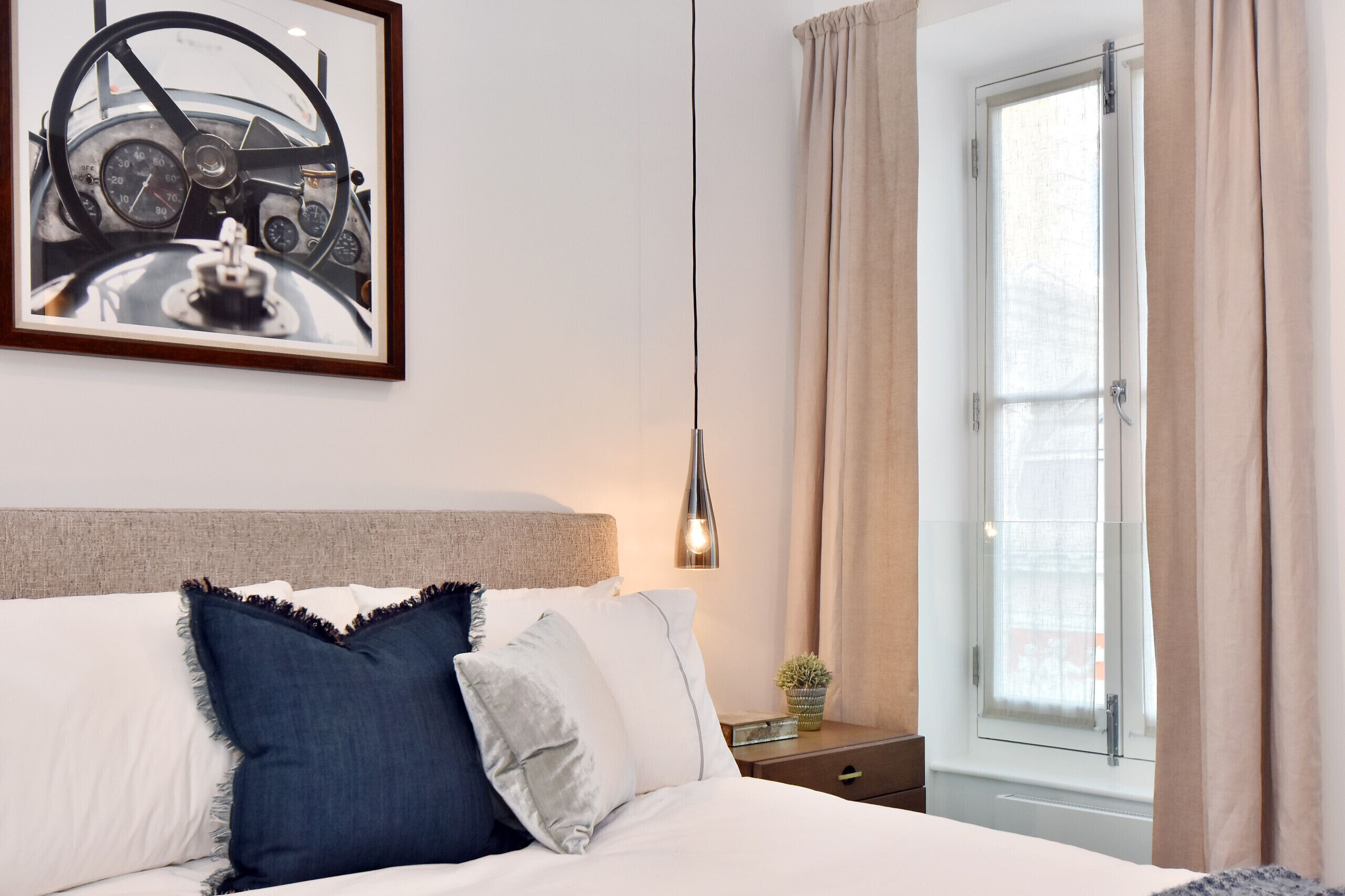 Een net opgemaakt bed met hoofdbord, een lijst met een afbeelding van een dashboard van een oldtimer, een bedlamp en een raam met gordijnen.