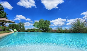 A clear blue pool with lounge chairs and umbrellas, surrounded by lush greenery and trees under a bright blue sky with clouds.