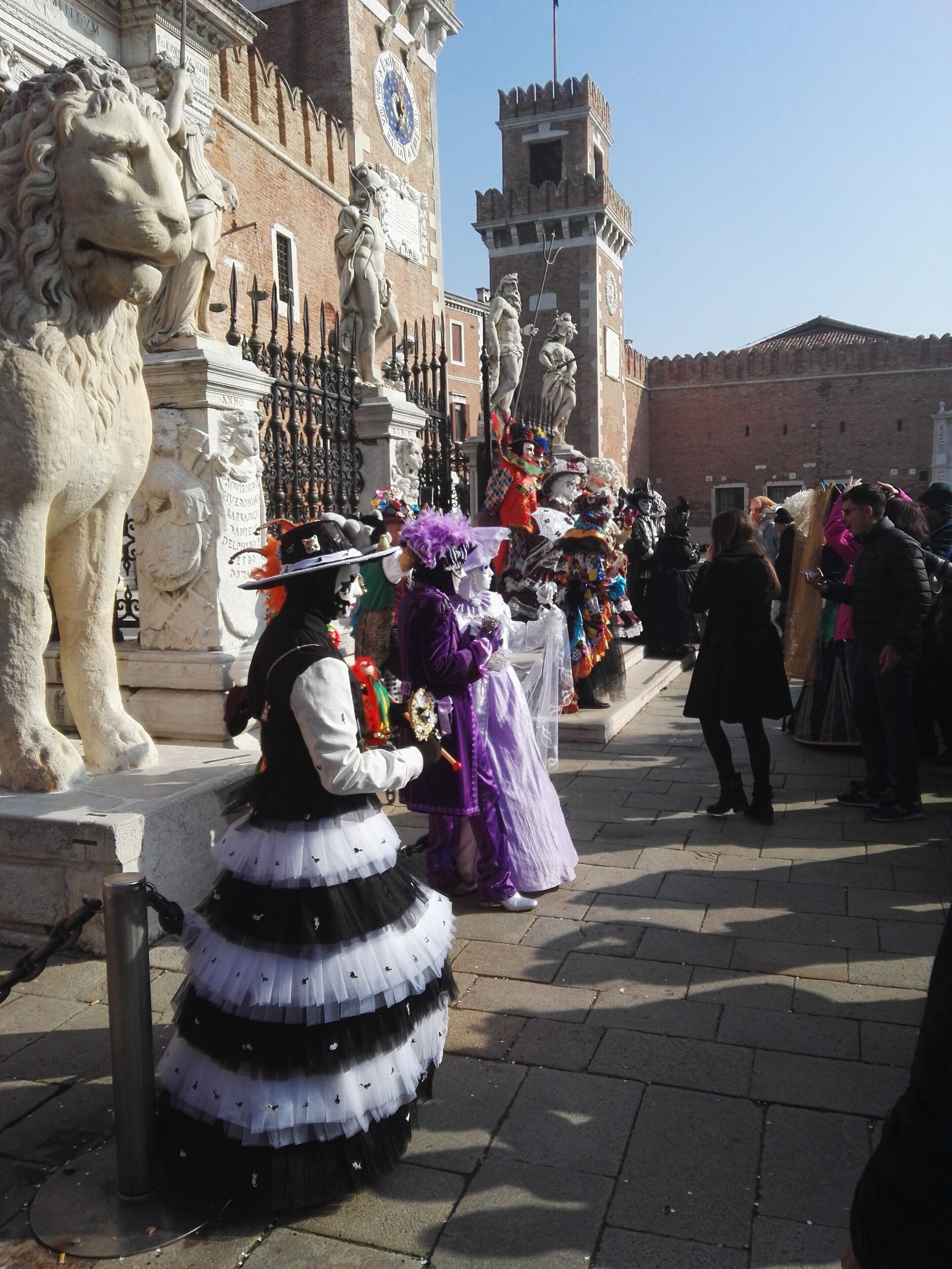 A historical square with statues, a clock tower, and people in period costumes.