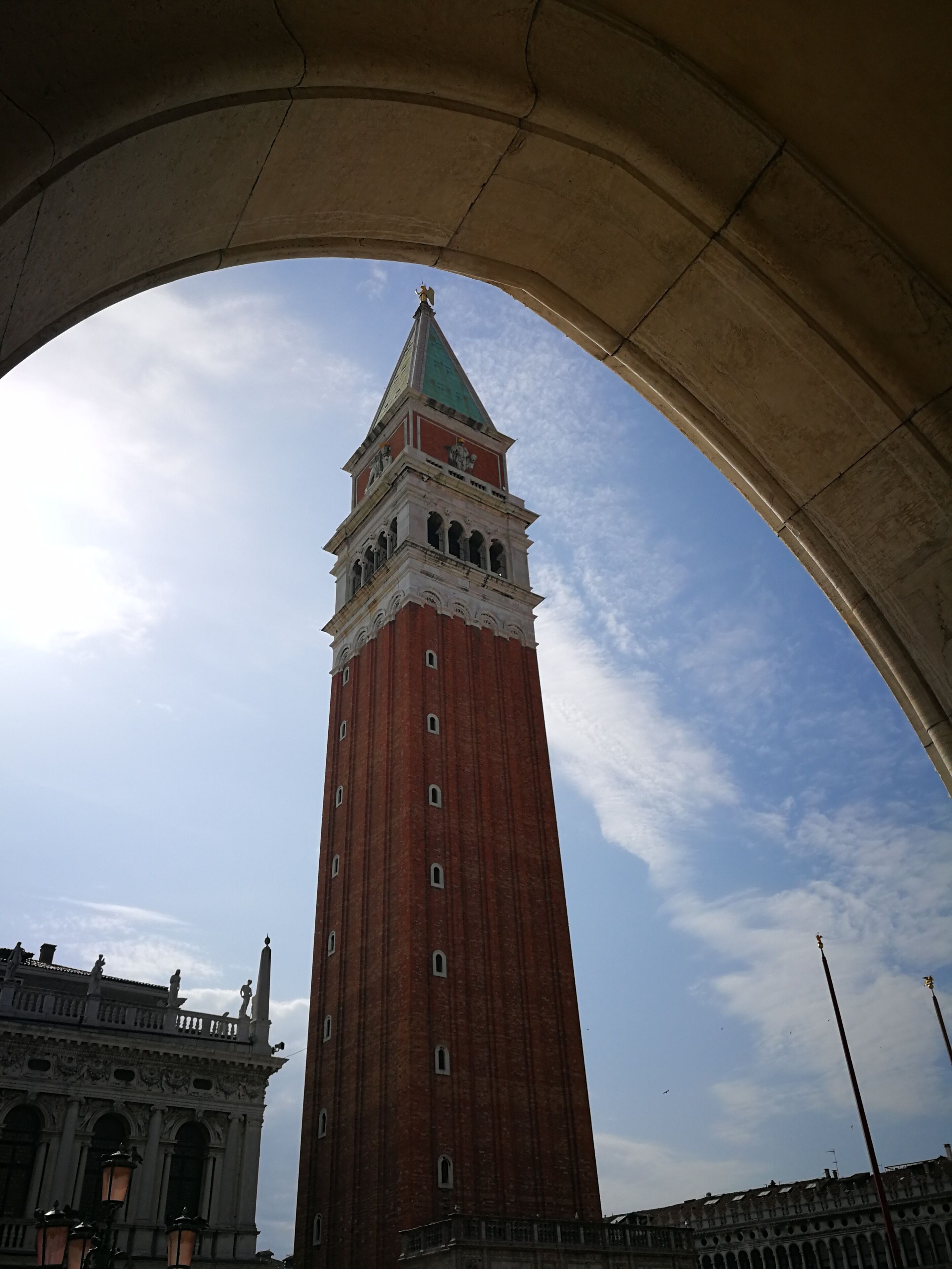 A tall red brick tower with a green roof and a clock, viewed through an archway.