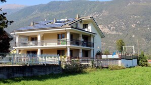 Una casa a due piani con un balcone, pannelli solari sul tetto e montagne sullo sfondo.