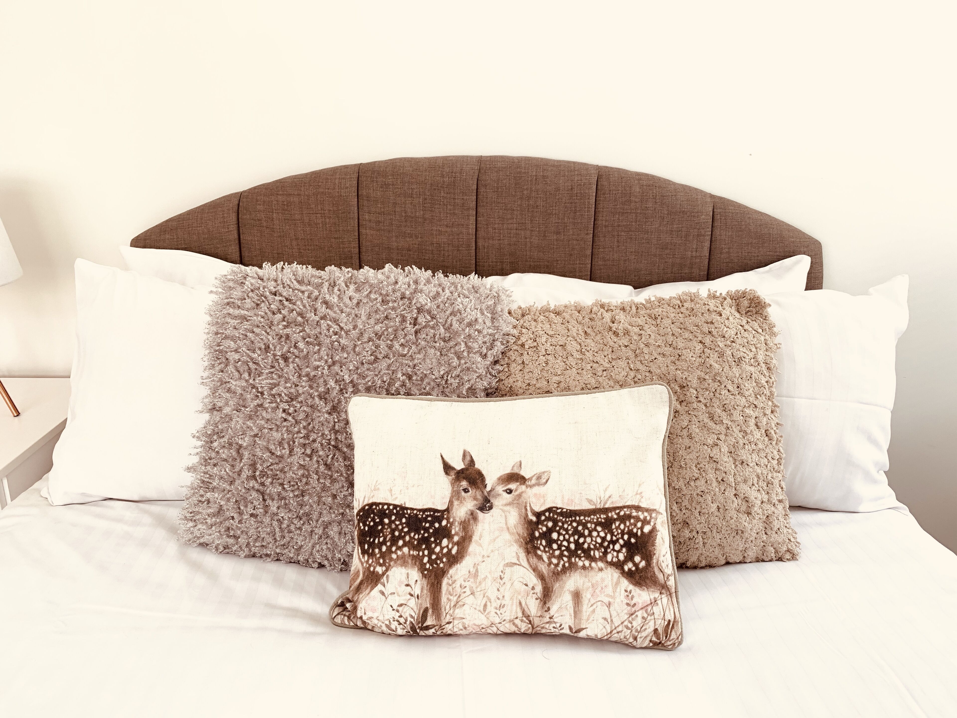 A bed with a brown upholstered headboard, multiple pillows including one with a deer design, and a fluffy grey throw blanket.