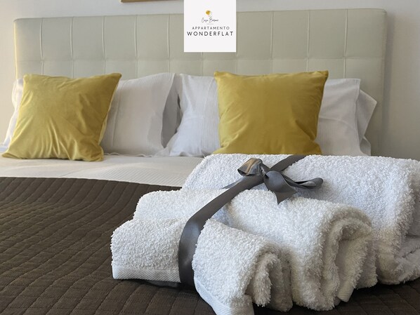 Una cama bien hecha con almohadas blancas y amarillas, una toalla blanca doblada con un lazo gris, y una colcha marrón con textura.