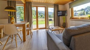 Ein modernes Interieur mit Holzbalkendecke, einem Essbereich mit Tisch und Stühlen, einer Couch, einem an der Wand befestigten Fernseher und einem großen Fenster mit Blick ins Freie.