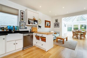 Una cocina moderna con armarios blancos, encimera de madera y lavavajillas empotrado. Hay un comedor con mesa y sillas de madera, y un ventanal que proporciona luz natural.