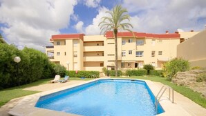 Une piscine rectangulaire avec un escalier, entourée d’une terrasse dallée. Au fond, on aperçoit un immeuble à plusieurs étages doté de balcons et d’un palmier.