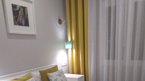 Una cama bien hecha con ropa de cama blanca y amarilla, una manta color esmeralda y una almohada con estampado floral. Hay una flor blanca sobre una toalla, y un cuadro en la pared.