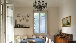 Een eetruimte met een grote tafel, witte stoelen, een kroonluchter, een raam met uitzicht en een schilderij aan de muur.