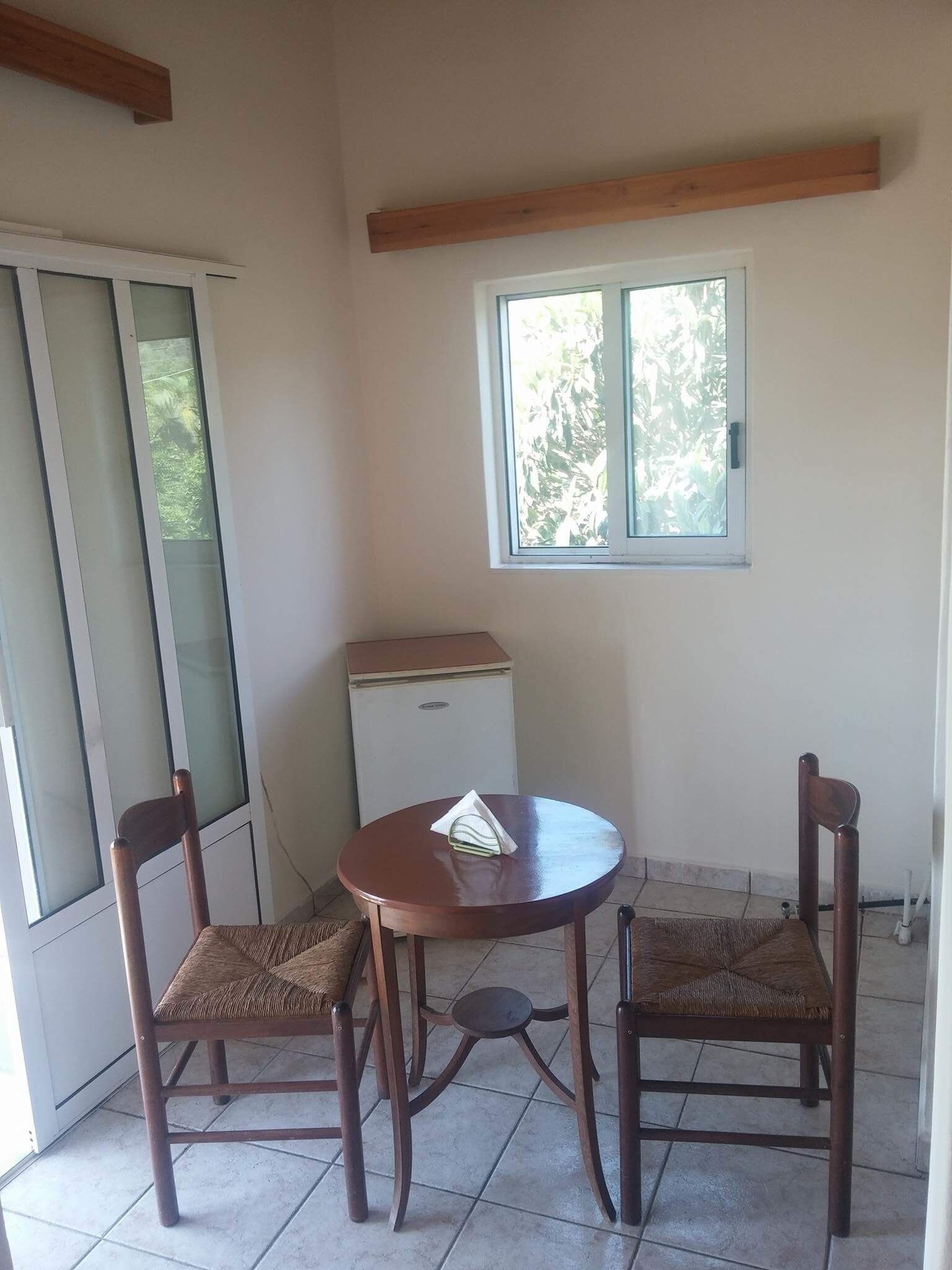 Une petite pièce avec une table ronde en bois et deux chaises, une fenêtre et un réfrigérateur.