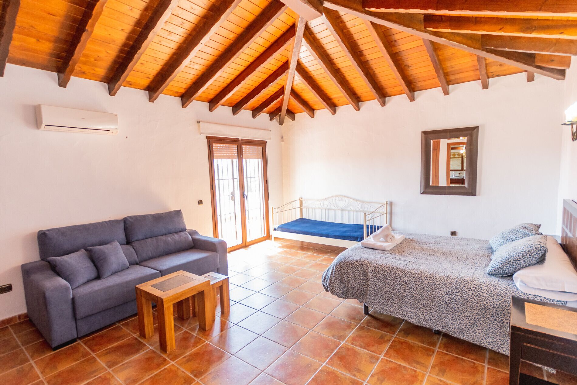 A cozy room with a wooden ceiling, a gray sofa, a bed with a patterned blanket, and a small wooden table.