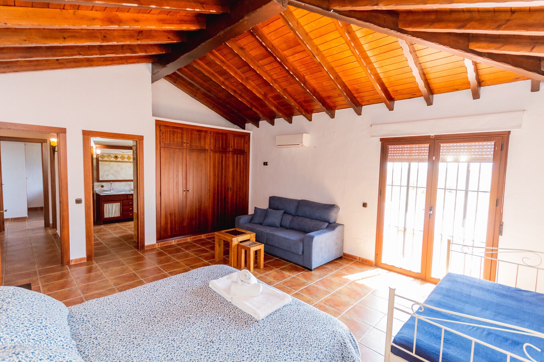 A spacious room with a wooden ceiling, a grey sofa, a bed with a blue and white patterned bedspread, and a small wooden table.
