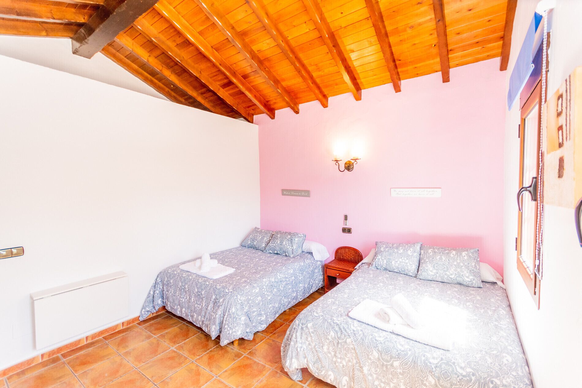 Un dormitorio con dos camas, techo de madera, paredes rosas y suelo de baldosas.