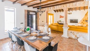 Un comedor con una mesa de madera dispuesta para seis personas, rodeada de sillas. Se observa un sofá amarillo al fondo y un pilar de piedra.