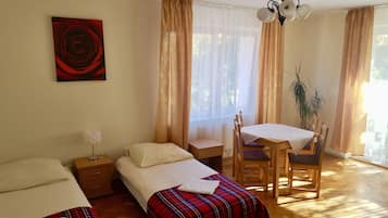 A bedroom with a bed, a bedside table, a small desk with a chair, a potted plant, and a window with curtains.