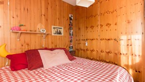 Ein Schlafzimmer mit karierter Bettdecke, Holzwänden, einem Regal mit Dekoration und einer Wandcollage mit Fotos.