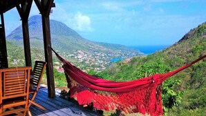A hammock on a wooden deck overlooking a coastal town with mountains in the background. - ❤T2 of standing, sea view, 2 minutes from beaches, quiet, nature, wifi, air conditioning (Les Anses-d'Arlet)