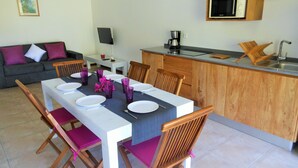 A dining area with a table set for four, wooden chairs, a kitchen with a microwave, and a sofa in the background. - ❤T2 of standing, sea view, 2 minutes from beaches, quiet, nature, wifi, air conditioning (Les Anses-d'Arlet)