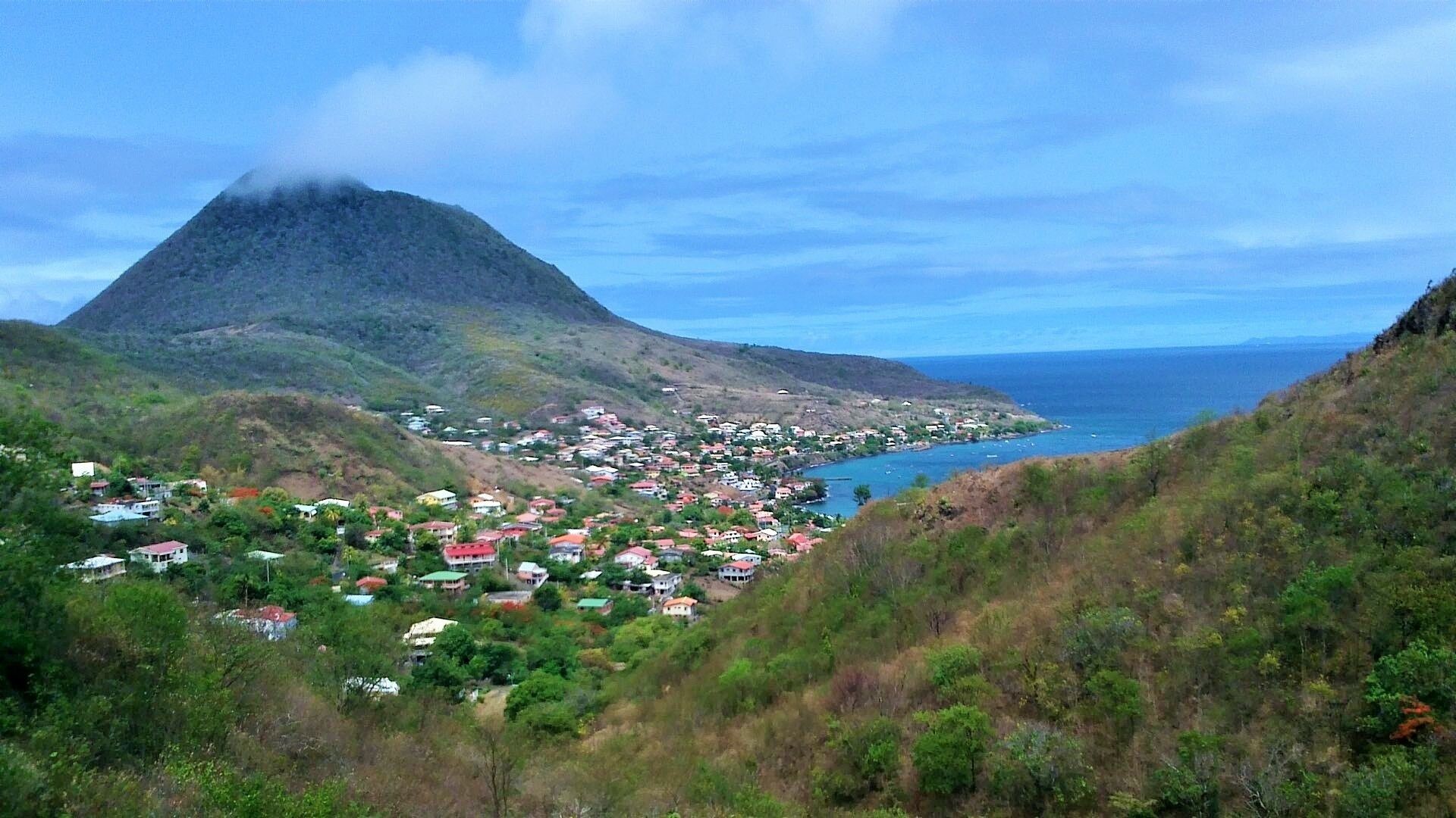 A coastal town with houses on a hillside, a mountain in the background, and the sea in the foreground.