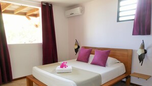 A bed with a wooden frame, white bedding, and two purple pillows. There is a small bedside table with a lamp, and an air conditioning unit on the wall. - ❤T2 of standing, sea view, 2 minutes from beaches, quiet, nature, wifi, air conditioning (Les Anses-d'Arlet)