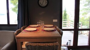 Una mesa de comedor dispuesta con platos y vasos, un reloj en la pared, y vistas a una terraza con un gato afuera.