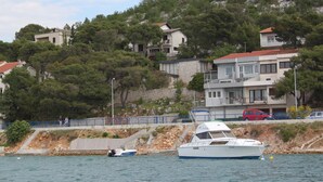 Zona costera con un yate blanco amarrado cerca de una costa rocosa, rodeada de edificios con balcones y un muro de piedra.
