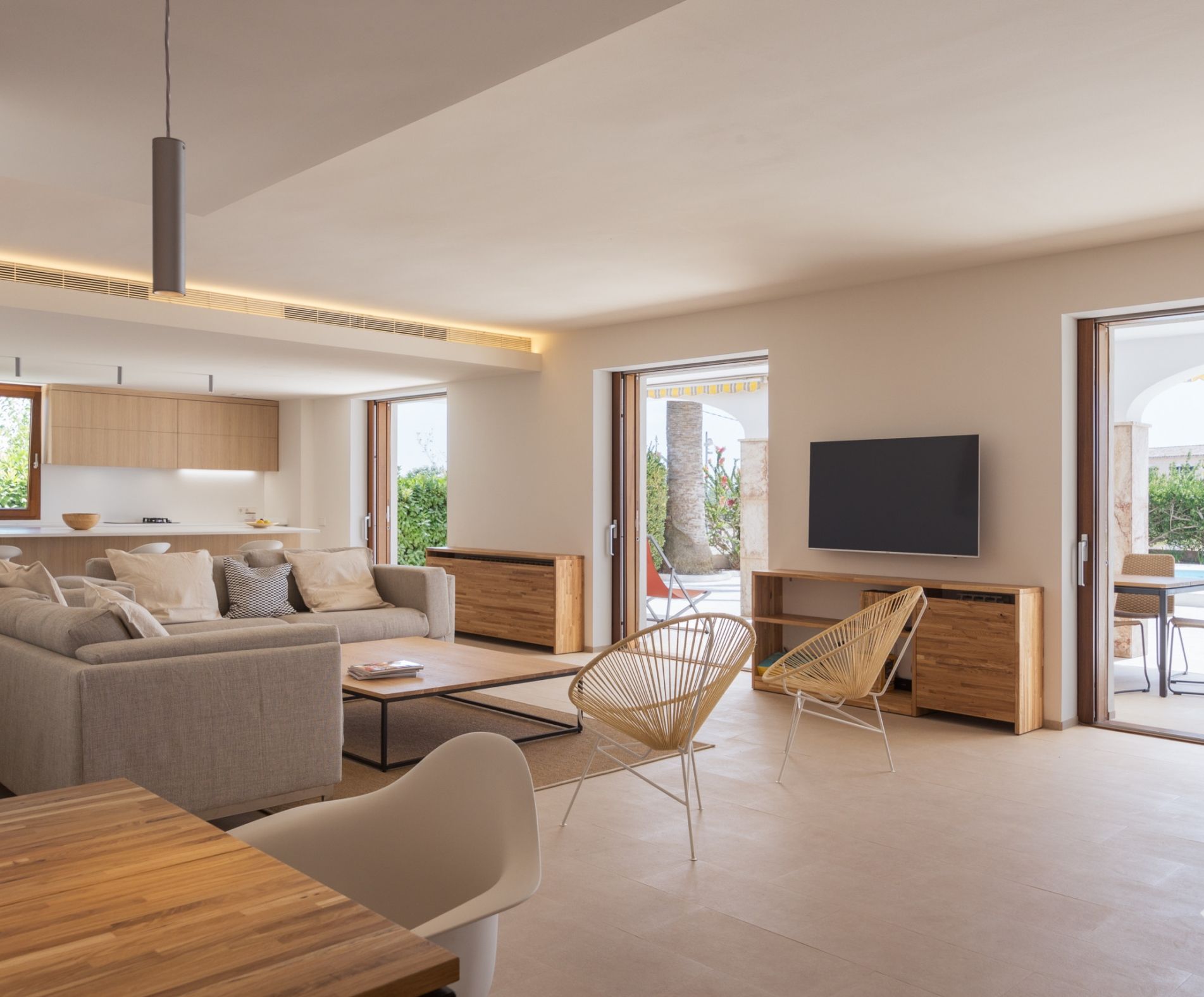 A modern living room with a sofa, coffee table, and chairs, a flat-screen TV mounted on the wall, and large windows offering a view of the outdoors.