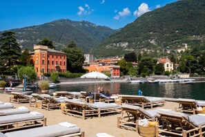 A beach with sun loungers and a view of a town and mountains in the background.