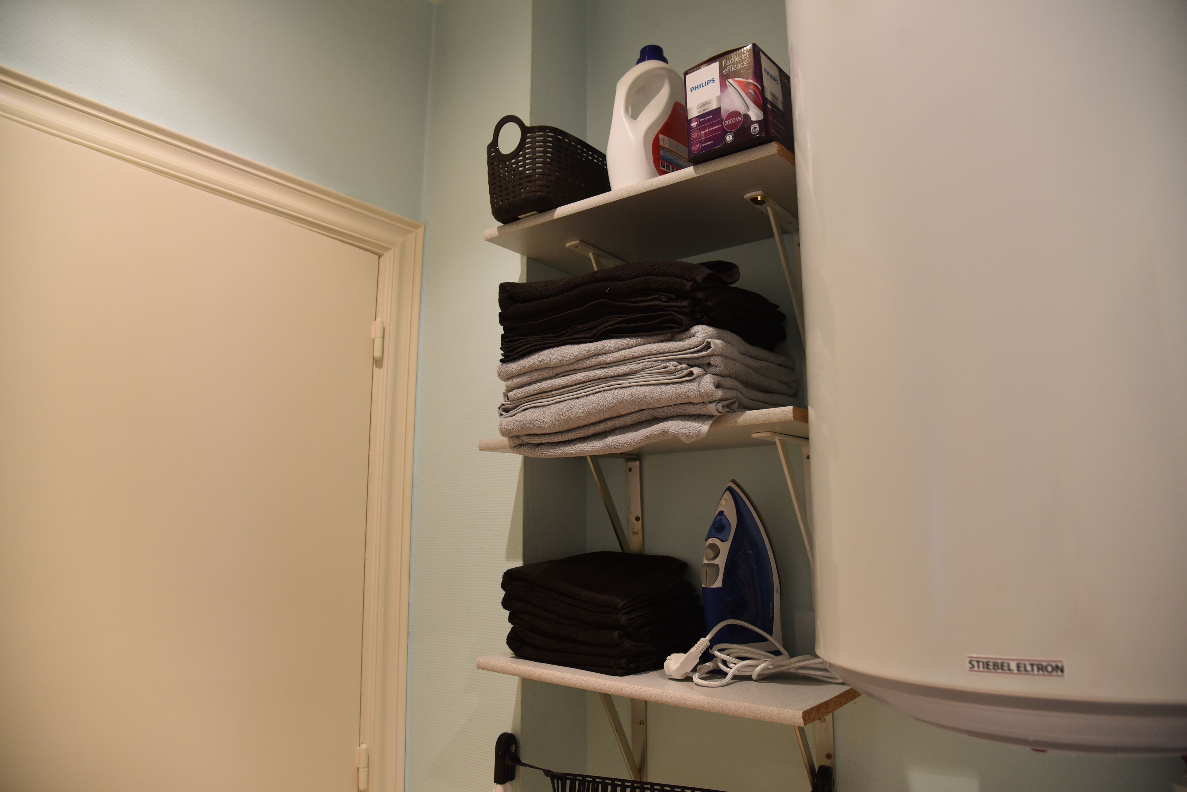 Un espace dédié à la lessive, avec des étagères où sont pliées des serviettes, un panier, une bouteille de lessive, un fer à repasser et une boîte de lessive.