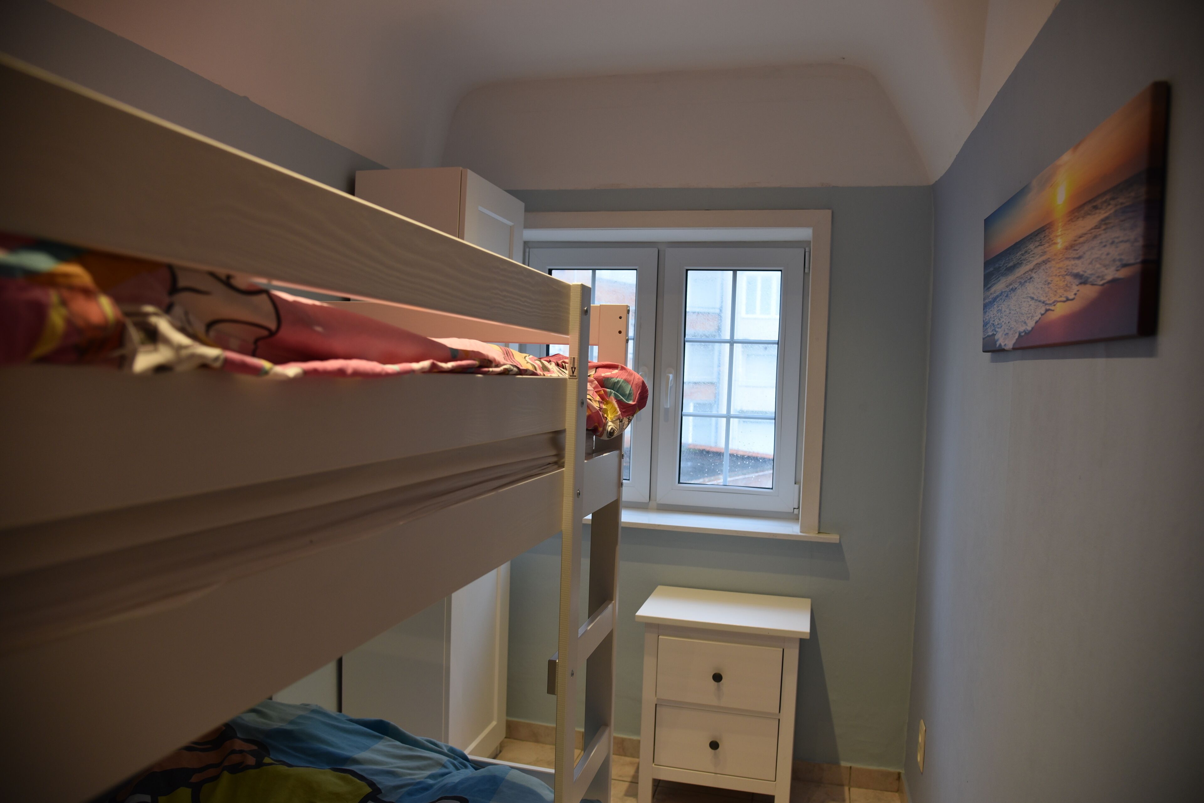 Une chambre avec des lits superposés, une petite table de chevet, une fenêtre et un tableau accroché au mur.