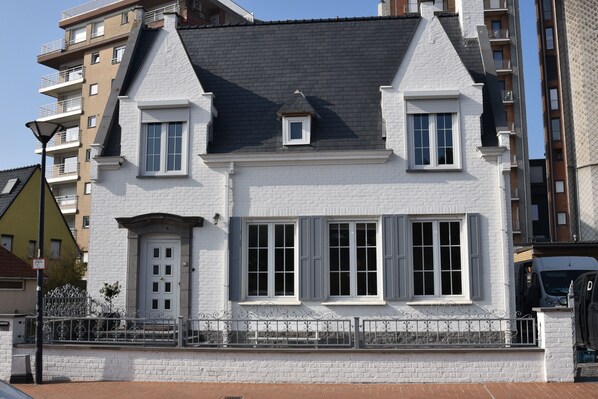 A white building with a gabled roof, two windows with shutters, and a small dormer window.