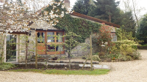 Une maison en pierre avec un porche en bois, entourée d’une allée gravillonnée et d’un jardin où poussent diverses plantes et fleurs.