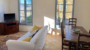 Un salon comprenant un canapé, un téléviseur posé sur un meuble en bois, un lustre et deux fenêtres donnant sur des arbres.