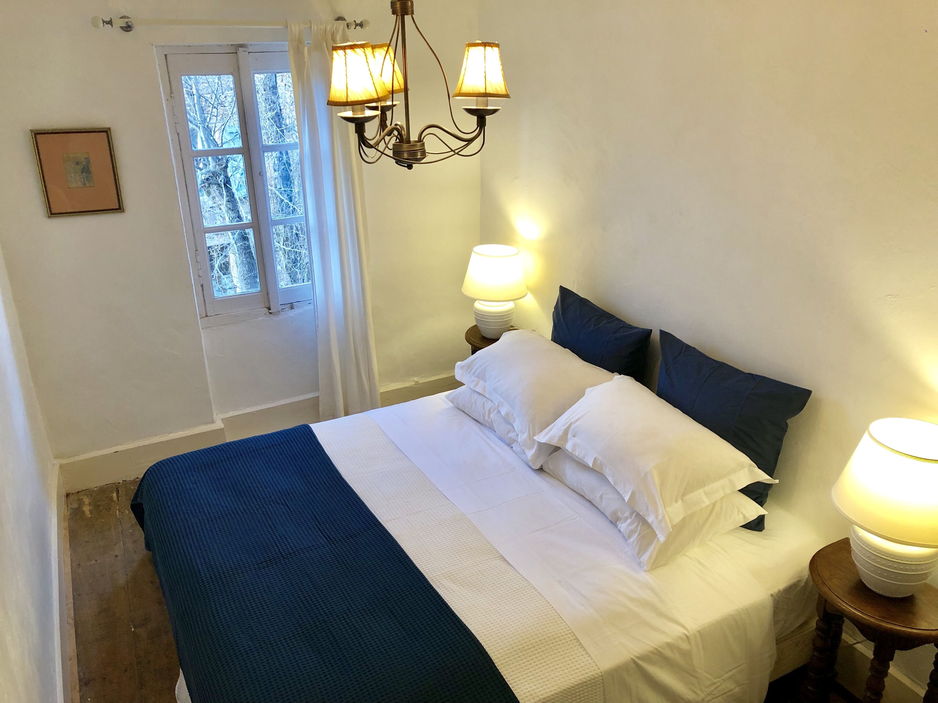 A bedroom with a bed, two bedside lamps, a window with curtains, and a picture on the wall.