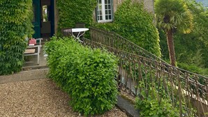 Un immeuble de deux étages avec une façade en pierre, un petit balcon agrémenté d’une table et de chaises, et une vue sur la verdure environnante.