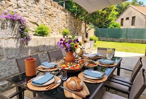 Un espace de restauration en plein air avec un mur en pierre, une table dressée avec des assiettes, des couverts, une miche de pain, un pichet et un saladier rempli de fruits.