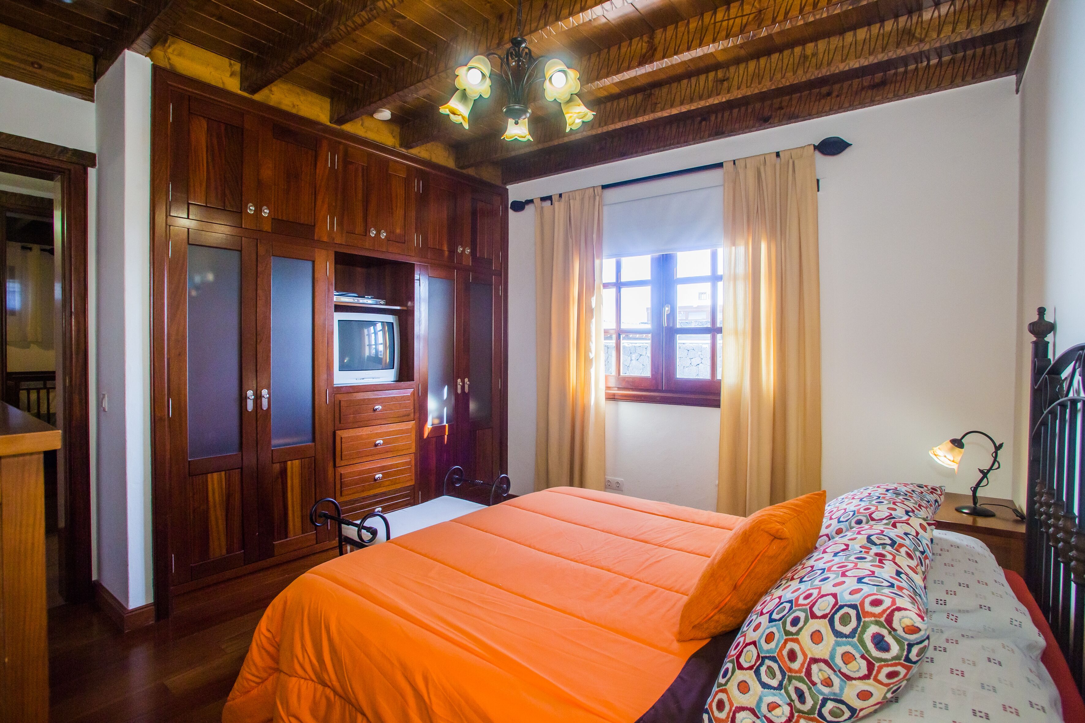 A bedroom with a wooden wardrobe, a bed with an orange bedspread, a patterned pillow, and a window with curtains.