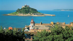 Un pueblo costero con un castillo imponente en una colina, un muelle que se adentra en el mar, y una iglesia con una aguja distintiva.